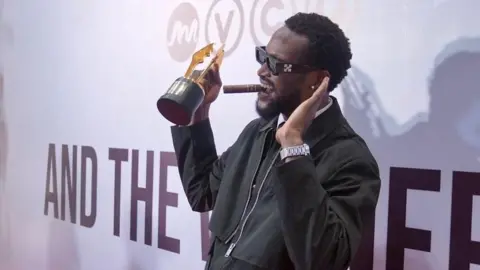 BBC/ MOBOLAJI OLATUNDE Bucci Franklin, in a black outfit and wearing shades, is on the stage with his award. 