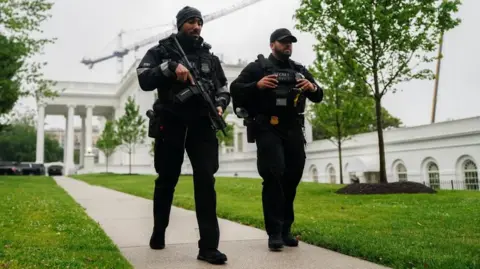 Reuters Two Secret Service agents in black uniforms and black hats walk alongside one another on a path outside the White House. One agent is carrying a large gun across his chest. 