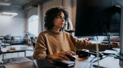 Getty Images Woman with dark curly hair, wearing tan polo neck jumper, works on a computer in an office 
