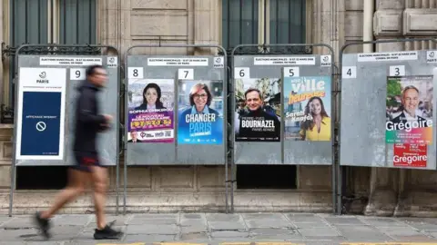 Reuters A man runs past electoral campaign panel boards with posters of Paris mayoral election candidates Sophia Chikirou of La France Insoumise (LFI- France Unbowed), right-wing Rachida Dati, Pierre-Yves Bournazel of the Horizons party, Sarah Knafo of the Reconquete party, and Emmanuel Gregoire of the Socialist party, ahead of the upcoming mayoral elections in Paris, France, March 6, 2026