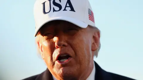 Reuters Trump pictured on 13 March. It is a close-up shot of his face in front of a blue sky. He wears a large white baseball cap with USA in large black letters on the front and a US flag on the side.