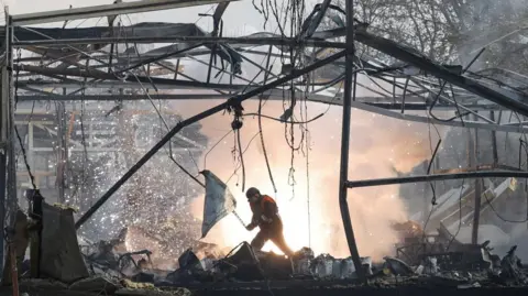 Reuters A rescue worker in the middle of rubble at a commercial building where only mangled metal frames remain; fire and smoke are shown in the background in the huge void betwen buldings