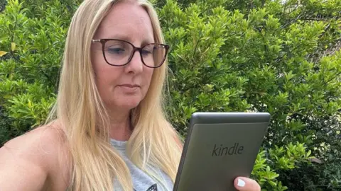 Kay Aaronricks Kay, wearing a grey vest top and glasses and stood in front of a green bush, holds her 5th Generation Kindle in front of her