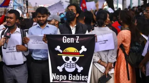 BBC A man in a black facemask holds a poster saying Gen Z Nepo Babies, with a skull and crossbones in the middle. Around him other protesters stand, holding printed signs. Flags can be seen being waved in the background