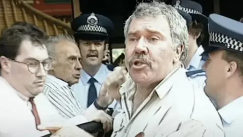 Seven Still from the video, showing Jack Karlson, wearing a beige shirt, surrounded by police and men in shirts trying to hussle him along as he speaks to the camera.