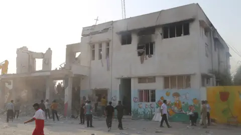 Reuters A still showing the aftermath of the strike on Minab. The exterior of the buidling is heavily damaged and people are seen outside inspecting it. 