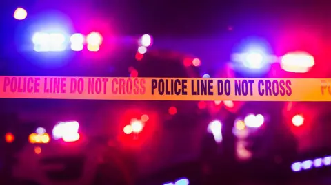 Getty Images Purple and pink flashing police lights are in rack focus in the distance with yellow police tape that says police line do not cross on it in the foreground.