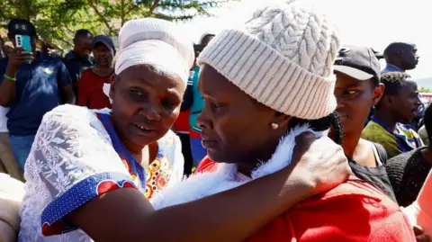 Reuters Two women - one in a white headscarf, the other in a white beanie hat, look worried as they embrace.