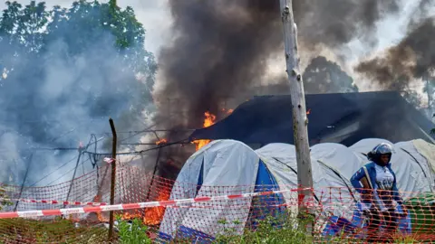 Reuters Fire and smoke at the of burnt tents on the grounds of Rwampara General Hospital. A police officer with a gun can be seen to the right of the shot in front of other makeshift tents.