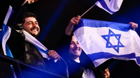 Hannibal Hanschke/EPA Two people, perched overlooking a metal railing, are smiling and celebrating. The man on the left has short black hair and a black goatee. He is draped in an Israeli flag, which is blue stripes, with a large stripe of white in the middle and a blue Star of David. The woman on the left is holding a small flag of Israel on a stick as well as a larger flag which she is waving in both hands. 