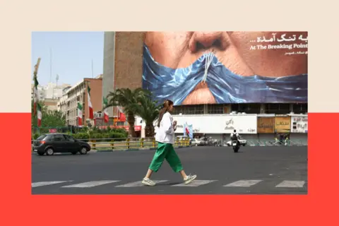 Reuters A woman wearing a white shirt and green trousers walks in front of a billboard showing the Strait of Hormuz as a gag stitched across Trump's mouth 