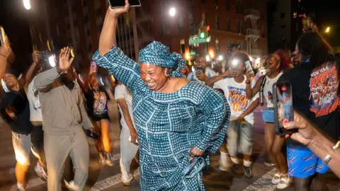 Getty Images A woman wearing a matching headscarf and dress raises her arm in the air as she dances on a zebra crossing in the street at night time. A crowd of other people dance around her, some raising their phones taking pictures. 