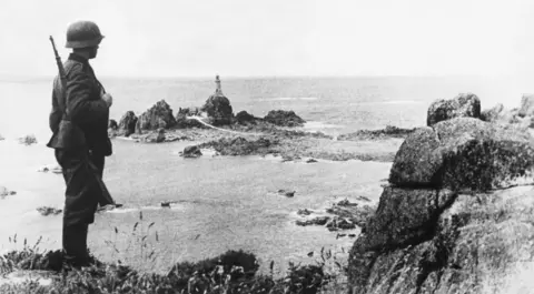 Atlantic-Press/Ullstein bild via Getty Images A German soldier stands on a rocky ledge, with a small rocky peninsula topped by a lighthouse in the background.
