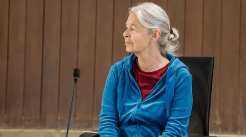 Pool/Getty Images A woman with greying hair sits in front of a microphone, wearing a blue zip-up sweatshirt and red undershirt