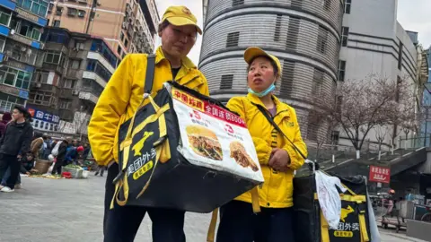 Getty Images Two food delivery personnel in China wearing yellow uniforms, walking in the middle of a city, carrying big bags with photos of a sandwich and fried chicken