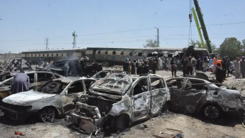 Getty Images Burnt-out cars are seen in front of a derailed train, while people stand around.