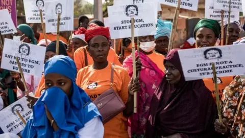 AFP via Getty Images Anti-Female Genital Mutilation (FGM) protesters hold placards outside the National Assembly in Banjul on march 18, 2024, during the debate between lawmakers on a highly controversial bill seeking to lift the ban on FGM