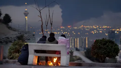 Anadolu via Getty Images Three Tehran residents sit on a bench overlooking smoke billowing in the distance at night on 8 March.
