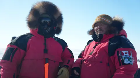 Eloise Alanna/BBC Two men -in the red, fur-trimmed parka uniforms of the Canadian Rangers. One has his face completly covered by shaded goggles, the other wears a hat and has his hood down. 