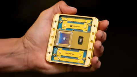 Microsoft A hand holding the Majorana 2 chip in its palm. Including its housing the chip appears to be about the size of a beer mat. It consists of a gold plated metal frame and sits on a dark blue-green board bearing its name. The chip itself is a small silvery square on a dark gunmetal blue square with very fine gold circuitry visible. 