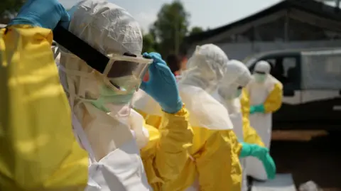 A man in yellow and white PPE, including eye protection and blue gloves. Three other people in similar gear can be seen behind him - Bunia.