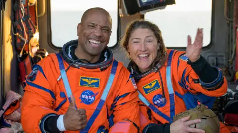 Reuters Victor Glover and Christina Koch smile in their orange space suits after returning to Earth. They are sat on a Navy MH-60 Seahawk, the interior of which is visible behind them.