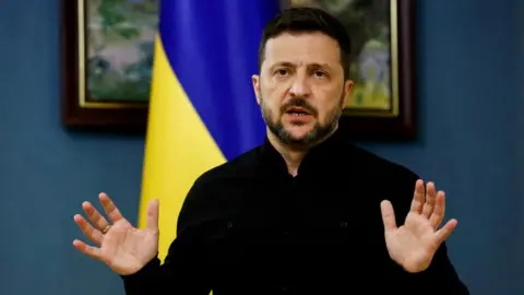 Reuters President Zelensky in his trademark black top holding his hands up in front of a Ukrainian glad and two paintings on a dark blue wall