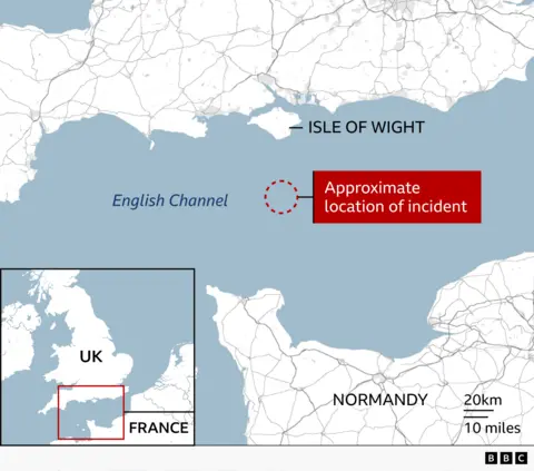Map of the central English Channel showing the south coast of England and the north coast of France. The Isle of Wight is labelled off England’s south coast, and Normandy is labelled along the French coastline. A dashed red circle in the sea between the two coasts marks the “approximate location of incident,” highlighted by a red box and arrow. An inset map in the lower left shows the wider UK and northern France region with the highlighted area. A scale bar indicates 20 km (10 miles), and a BBC logo appears in the lower corner.