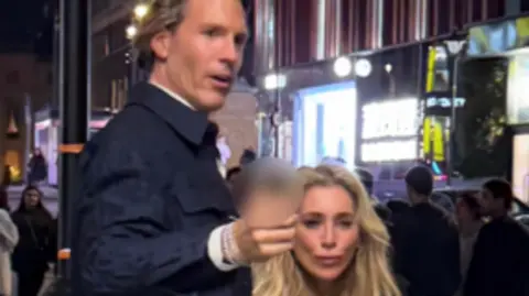 British Transport Police A CCTV image showing a tall man with brown hair wearing a smart dark blue jacket, gesturing forward. He is with a woman with long, blonde hair 