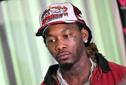 Getty Images Offset is a man, he has brown eyes and is wearing a white and red cap. He has a diamond stud earring and is wearing a large silver chain. He is wearing a red jacket with a black collar. He has a tattoo on his cheek below his eye of a cross and tattoos on his neck. 