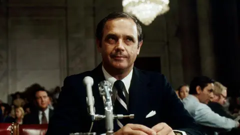Getty Images Alexander Butterfield sits before the Senate Watergate Committee wearing a black suit, with a microphone in front of him.