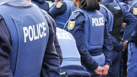 Gallo Images via Getty Images Close up of the backs of police officers standing in formation. The are in uniform and vests with police written on them in white lettering.