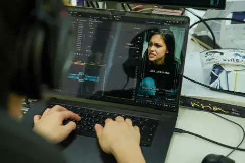 BBC/Hosu Lee A woman's face is seen on a laptop screen in editing software, with the computer user's head and hands visible.