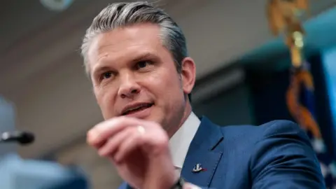 Getty Images Head and shoulders pic of Pete Hegseth at the podium of the Pentagon briefing room. He's wearing a well-tailored petrol blue suit with a blue and pink tie on a white shirt. His left hand is raised and his thumb and index finger joined as if emphasising a point