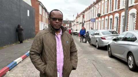 PA Media A man standing in a street lined with terraced houses. He is wearing a brown anorak and pink shirt and wearing dark-lensed glasses. He is balding, and has some hair towards the back of his head. On the right side there is a line of cars parked in front of the houses. On the left the kerbstones are painted red, white and blue. It is an overcast day. 