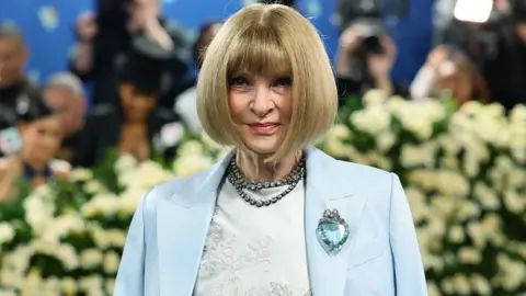 Getty Images Anna Wintour, with signature sharp blonde bob and fringe, wears a pale blue outfit with a matching jacket and jewelled brooch - flowers and foliage can be seen behind her 