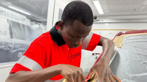 A young man wearing a hi-vis uniform tapes down plastic on a car windscreen in a workshop