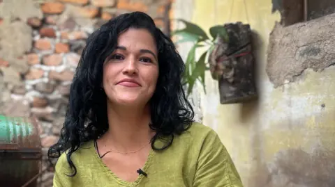 BBC Indira Martínez, who has long black curly hair, smiles at the camera. She is sitting on a patio, the paint on the wall is peeling and a metal oil drum can be seen in the background. She is wearing a lime-green top and a thin chain around her neck.