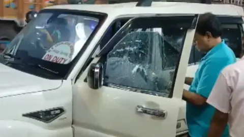 ANI Two men stand near the white car in which Chandranath Rath was travelling. The car window on the left side is open, and the glass is cracked