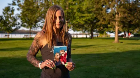 AFP via Getty Images Kim Prosser, wearing a brown tank top, stands in a park, looks towards the camera as she holds up a photo showing her with her son.