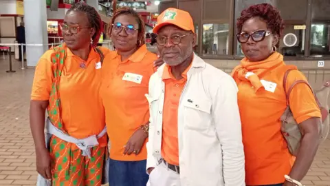 Julien Kouadio Adonis A man wearing an orange shirt and baseball cap and a white jacket looks at the camera. He has glasses and short facial hair. Three women stand with him, also wearing orange polo shits. 