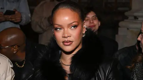 Getty Images Rihanna looks to the side with her hair slicked back. She wears several earrings and a black fur collared jacket at the AWGE fashion show as part of RTW Fall 2026 on 13 February 2026 in New York, New York. 