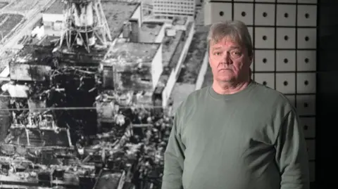 Nikolai Solovyov, chief engineer in turbine hall at time of accident, pictured in front of an image of the destroyed reactor, at the Slavutych museum. He has grey hair and is wearing a grey-green sweatshirt, and is looking at the camera with a serious expression.