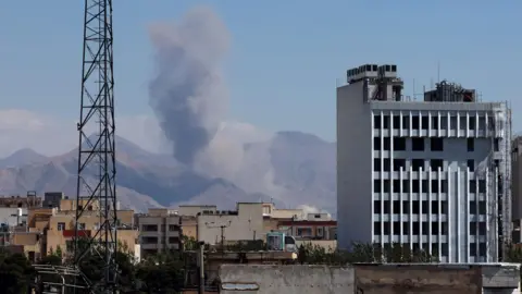 ABEDIN TAHERKENAREH/EPA/Shutterstock Smoke rises in the mountains in the distance as we look at building tops from far away in Iran as well as a metal tower.