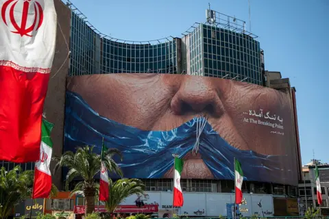 Getty Images A poster in Tehran depicts the mouth of U.S. President Donald Trump, gagged by a blue ribbon in the shape of the Strait of Hormuz, on May 9, 2026 in Tehran, Iran