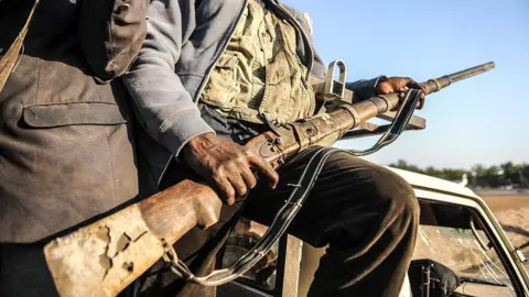 Getty Images A man sitting on the back of a car holding a gun. All that is visible of him is his torso, lap, arms and hands