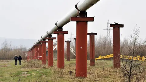 Bloomberg via Getty Images Men stand near the Druzhba crude oil pipeline near Stryi, western Ukraine, in 2009. The pipeline is situated on large orange pillars in a field.