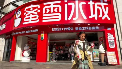Getty Images A shop of Mixue, a Chinese bubble tea brand in Chongqing, China in April 2026. In the foreground a girl walks away from the shop holding two sundaes and a drink.