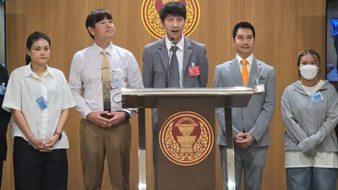 Supplied by Juraluk Kunaruk Juraluk Kunaruk (first from right), standing before the Thai parliament as MP Nitikorn Bunyakulcharoen (centre) addresses lawmakers on the spate of scams around BTS concert tickets