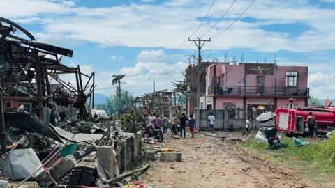 Shwe Phee Myay A charred wrecked building overlooks a dirt track on a sunny afternoon, with people standing around in the distance. 
